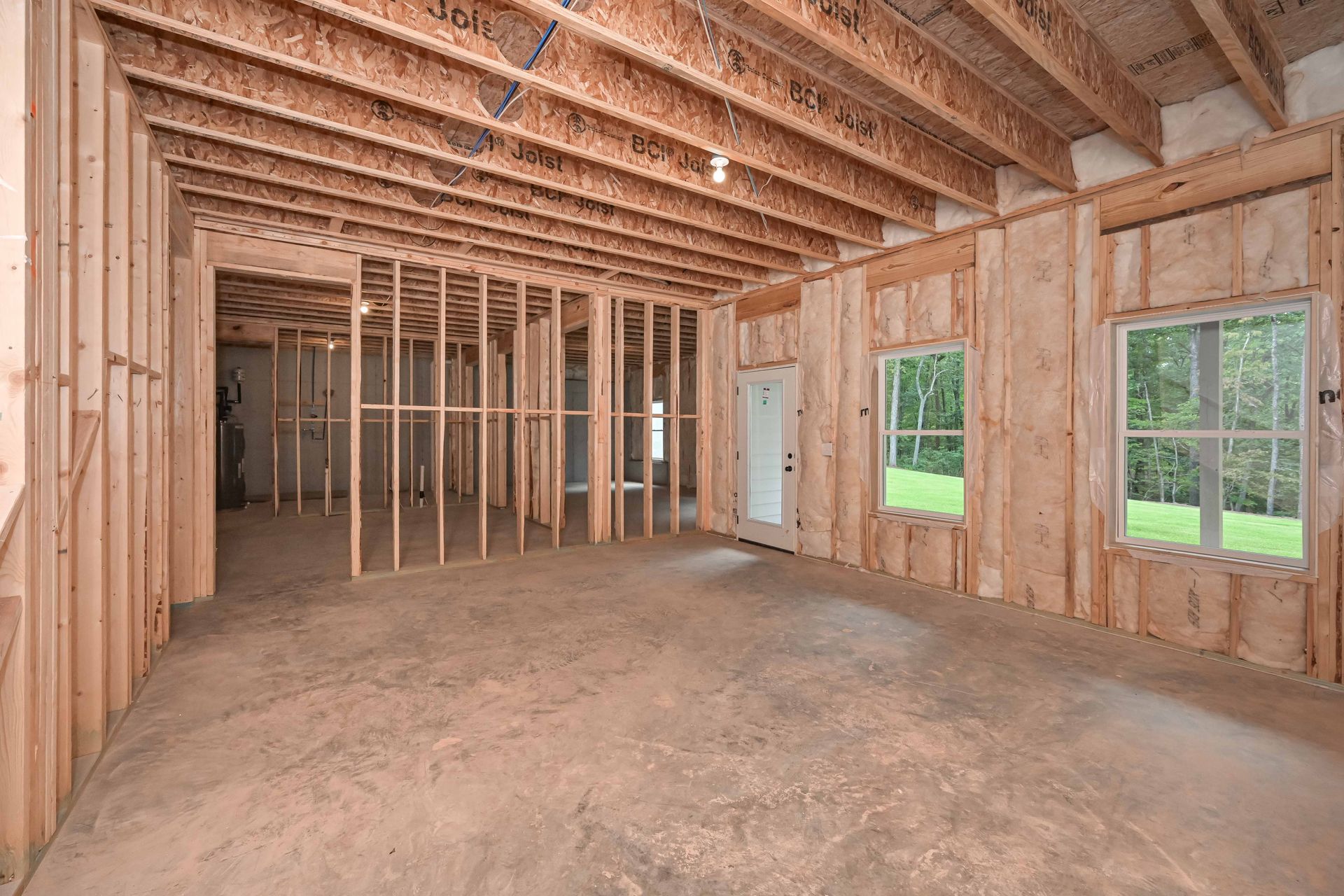 Interior of a house under construction; exposed wood framing for walls, ceiling, and floor; two windows and a door visible.