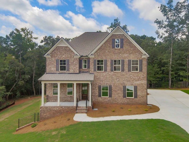 Two-story brick house with brown shutters, porch, and a driveway, set amidst a grassy lawn and trees.