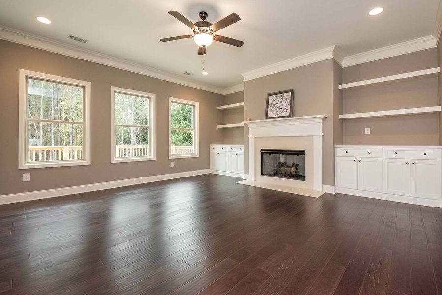 Empty living room with fireplace, built-in shelves, and dark hardwood floors. Three windows and a ceiling fan.