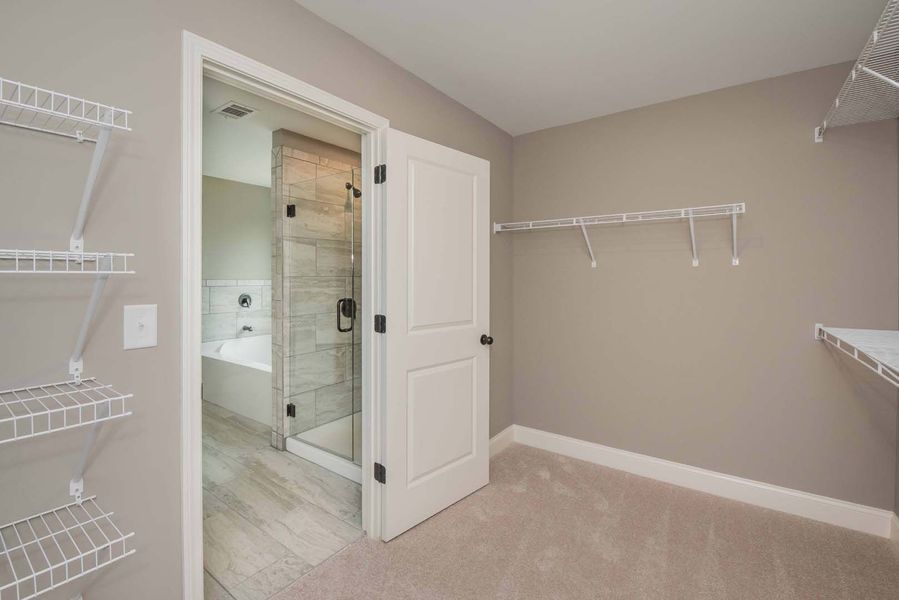 Walk-in closet with white wire shelving, beige walls, and a door to a bathroom with a tub and shower.