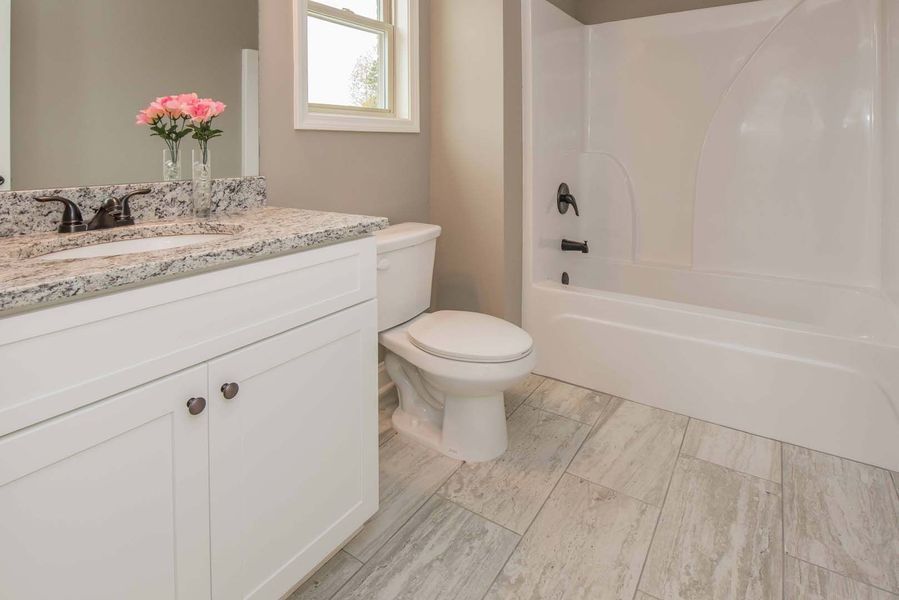 A modern bathroom with white fixtures, gray floor, granite countertop, and a window.