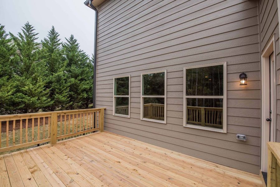 Wooden deck with railing, three windows, and a light fixture on a house with gray siding; forest in the background.