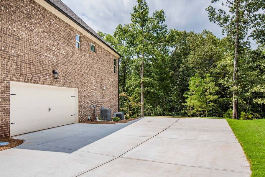 Brick house with concrete driveway, garage door, and grassy yard. Trees in the background.