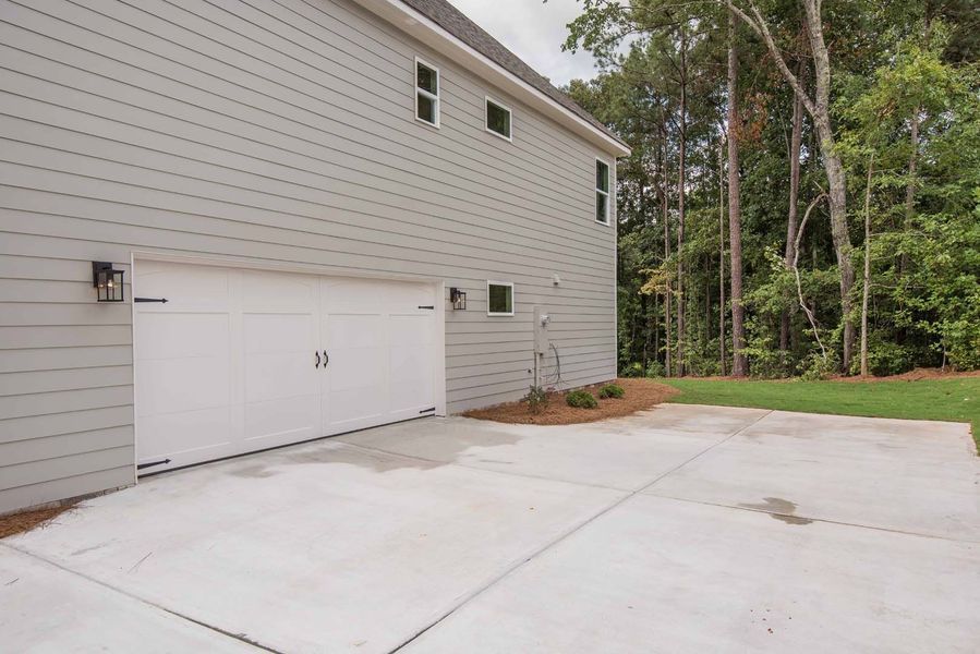 Side of a two-story gray house with a white garage door and concrete driveway. Lush trees in the background.