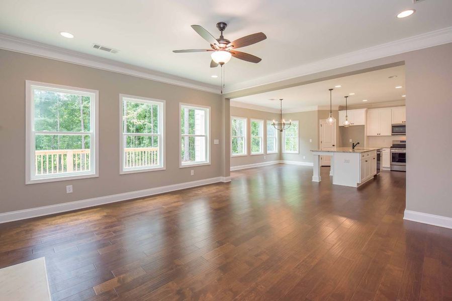 Empty, open-concept living room with hardwood floors, windows, and an adjacent kitchen. Neutral walls and white trim.