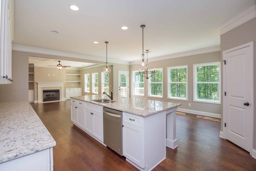 A kitchen with a white island, granite countertop, and stainless steel dishwasher, facing a living area with fireplace.