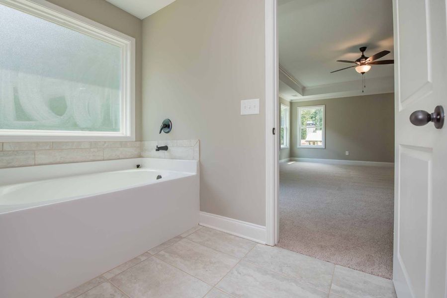 Bathroom with white tub, window, and view into bedroom with carpet, ceiling fan.