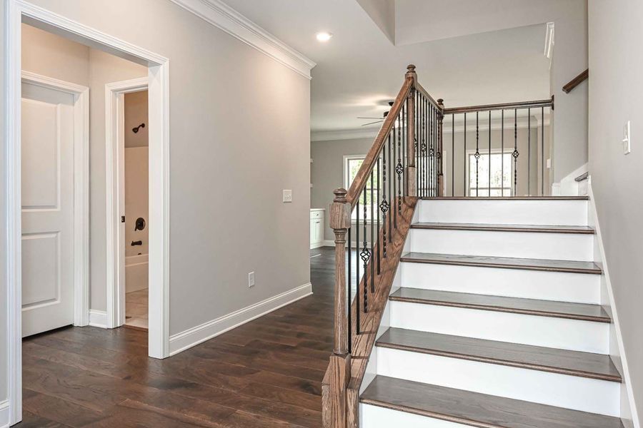 Hallway with staircase, gray walls, dark wood floor, white trim, open doorway to a bathroom.
