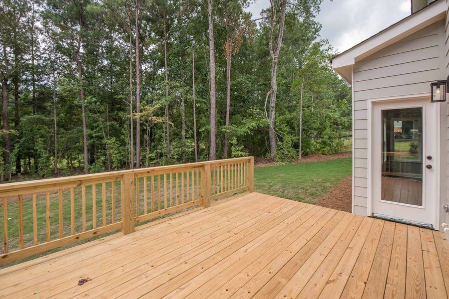 Wooden deck attached to a light-colored house with a glass door, overlooking a grassy area and tall trees.