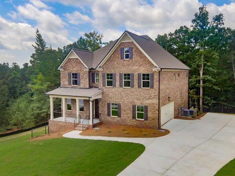 Two-story brick house with covered porch and driveway, surrounded by green grass and trees.