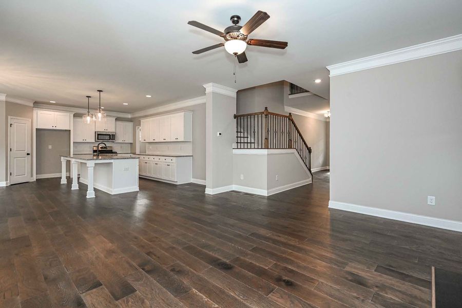 Spacious, empty living room with hardwood floors, connected to a white kitchen and staircase.