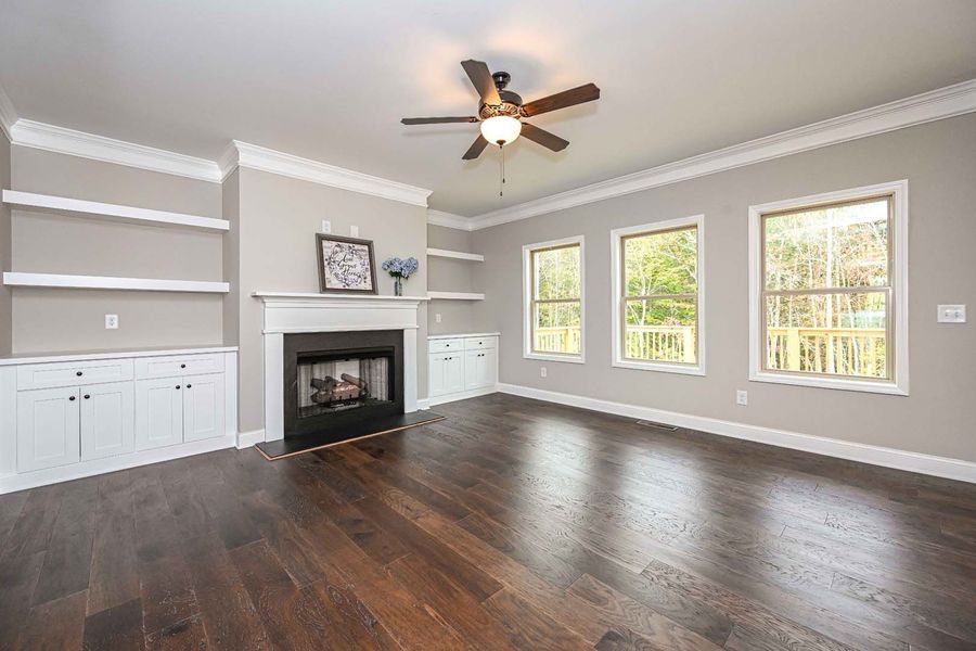Living room with fireplace, built-in shelves, hardwood floor, and windows. Neutral walls, white trim, ceiling fan.