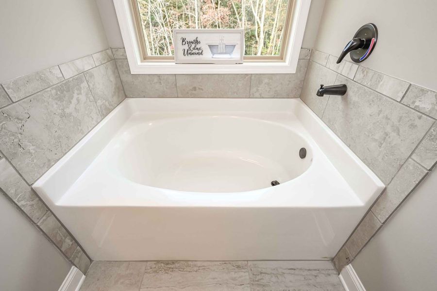 White built-in bathtub with black fixtures, under a window, surrounded by gray tiled walls.