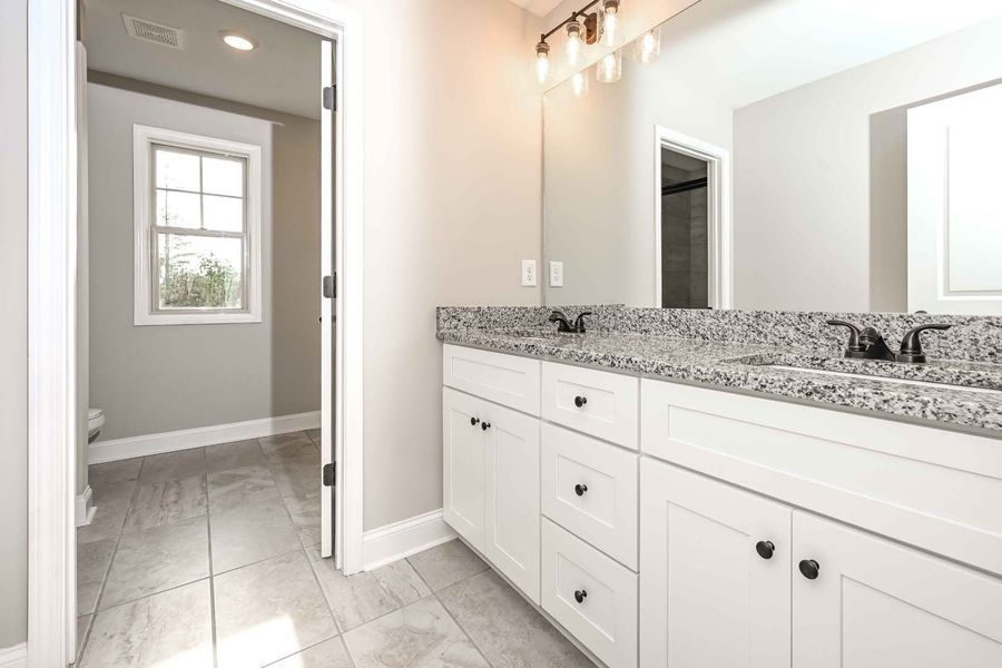 Bathroom with white cabinets, granite countertop, and a large mirror. A doorway leads to a window.