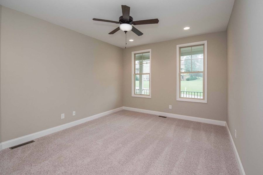 Empty bedroom with beige walls, carpet, two windows, and a ceiling fan.