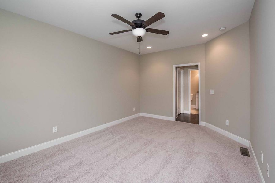 Empty bedroom with beige walls, gray carpet, and ceiling fan. Doorway leads to another room.