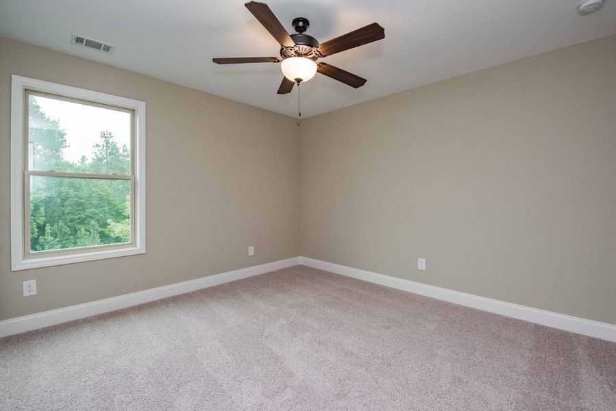 Empty bedroom with beige walls, carpet, and a ceiling fan. Window with outdoor view.