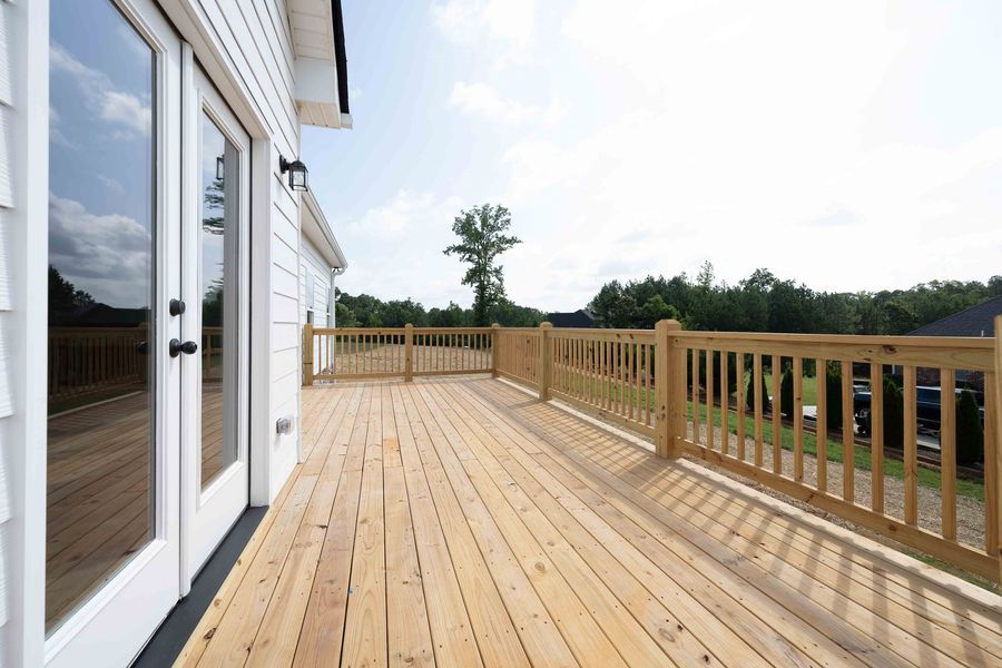 Wooden deck with railing, connected to a white house with glass doors, overlooking a green landscape.