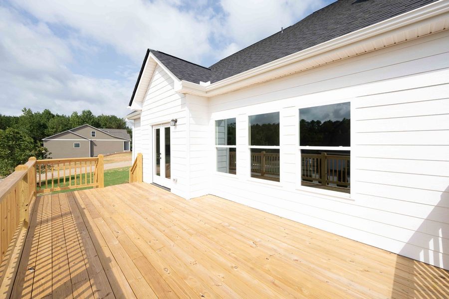 Wooden deck attached to a white house with black roof and three windows. Sunny day.