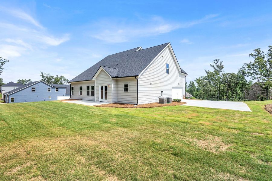 Backyard of a light-colored house with a gray roof and concrete patio, on a grassy lawn under a blue sky.