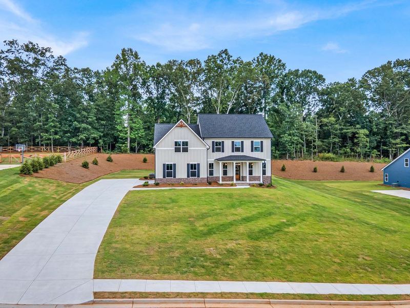 Two-story house with light gray siding, black roof, and a long concrete driveway on a grassy hill.