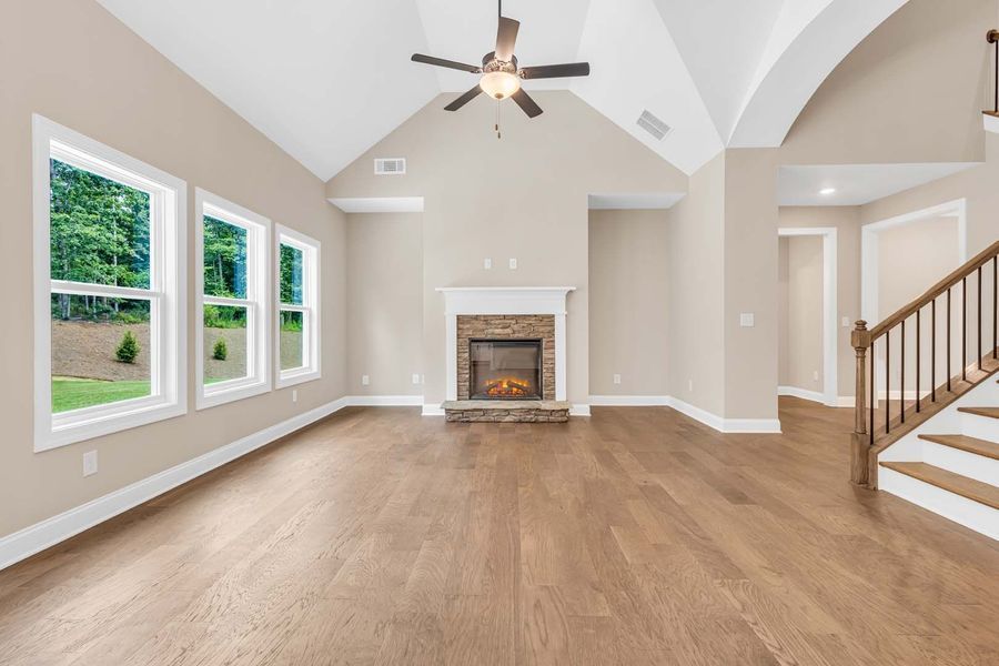 Empty living room with fireplace, windows, hardwood floors, neutral walls, and staircase.