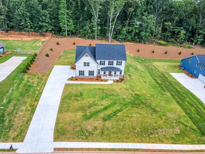 Two-story white house with blue shutters and a long driveway on a green lawn near a forest.