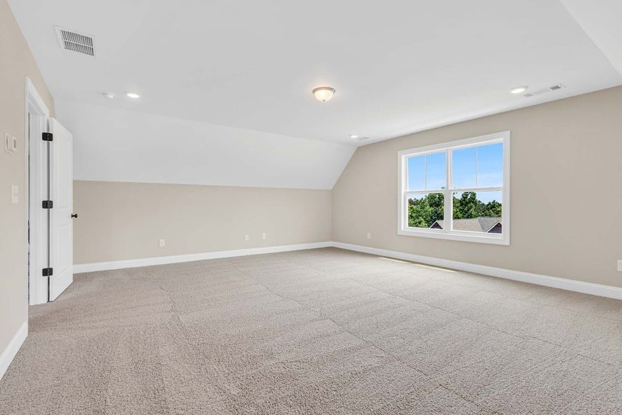 Empty, light-filled room with angled ceiling, carpeted floor, window, and closed door.