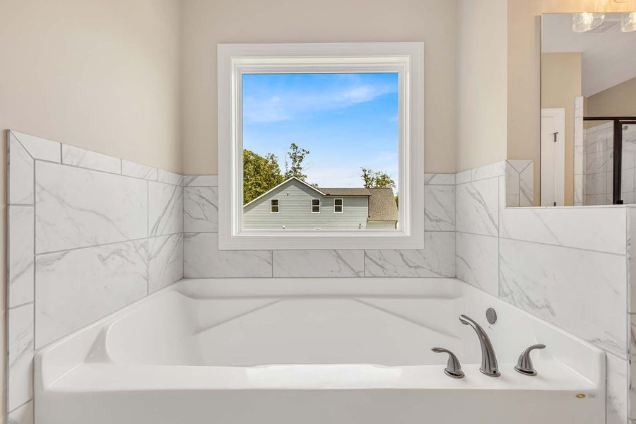 White bathtub with marble tile, beneath a window with a view of a house and blue sky.