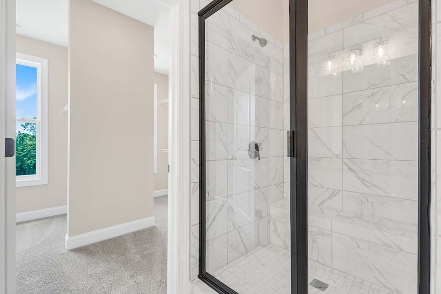 Bathroom with marble shower, black frame, and neutral walls; a hallway and window with trees in the background.