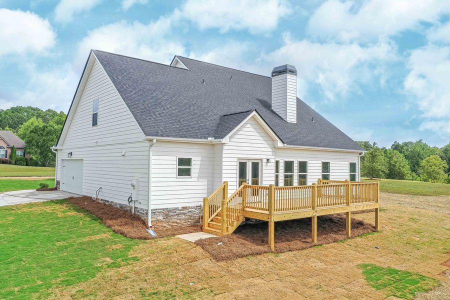 White house with a wooden deck, surrounded by grass and trees under a cloudy sky.