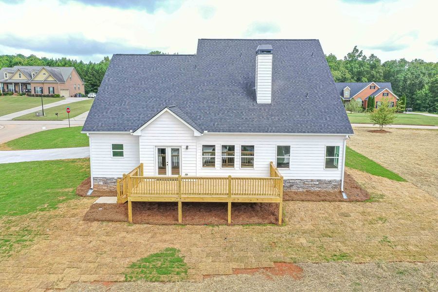 White house with wooden deck, set in a grassy yard. Other houses are visible in the distance.