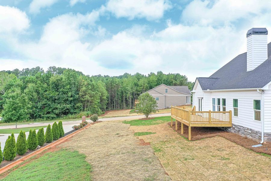 Backyard of a white house with a wooden deck, grassy lawn, and trees under a cloudy sky.