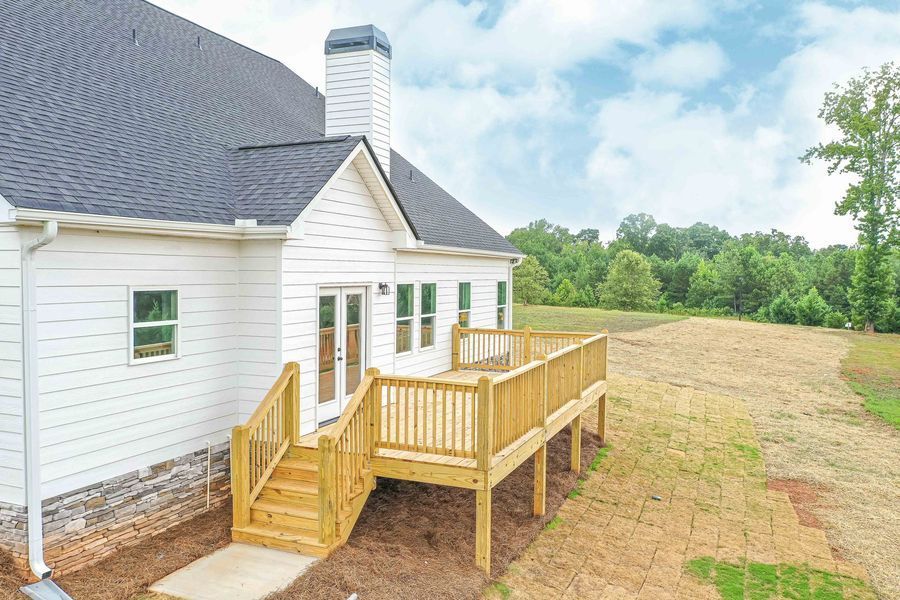 White house with wooden deck and stairs, surrounded by grassy field and trees.