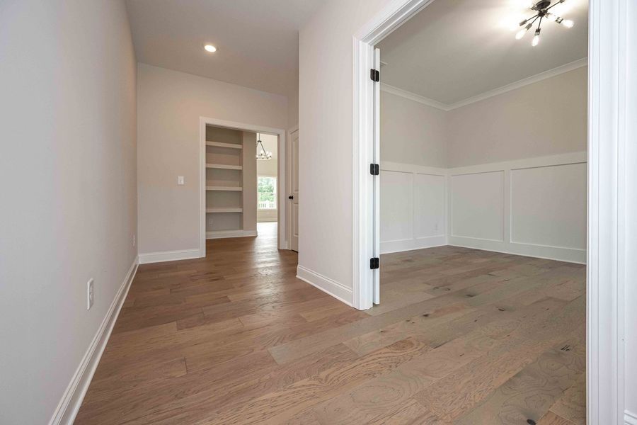 Hallway with wood floors, doorway to a room, shelves, and white walls.