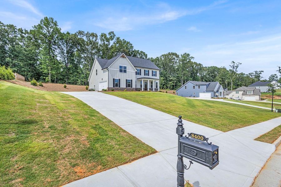 Gray house with long driveway, mailbox, and surrounding grass and trees.