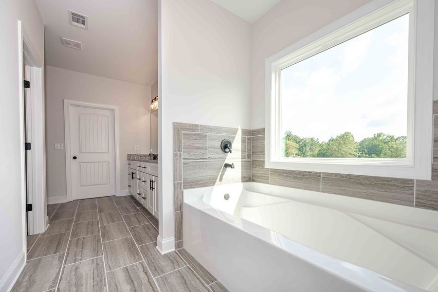 Bathroom with a white tub, window, and gray tile flooring. A doorway leads to a vanity.