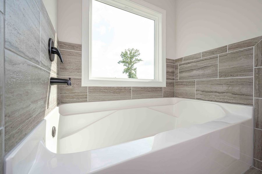 White bathtub with gray tile surround, black faucet, and window view of tree.