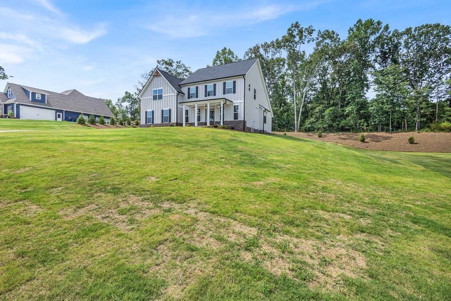 Two-story light gray house on a grassy hill with blue sky and trees.