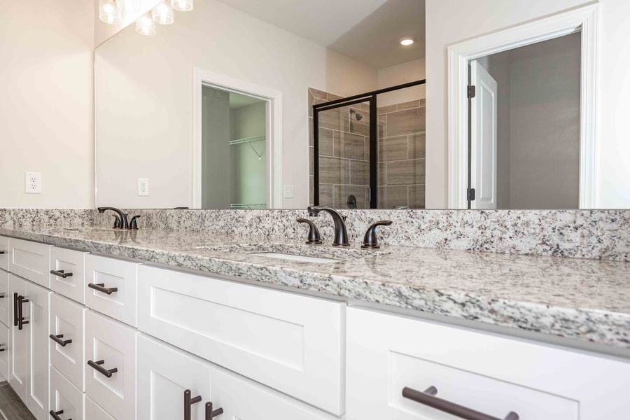 White bathroom vanity with granite countertop, black faucets, and a large mirror reflecting a shower and door.