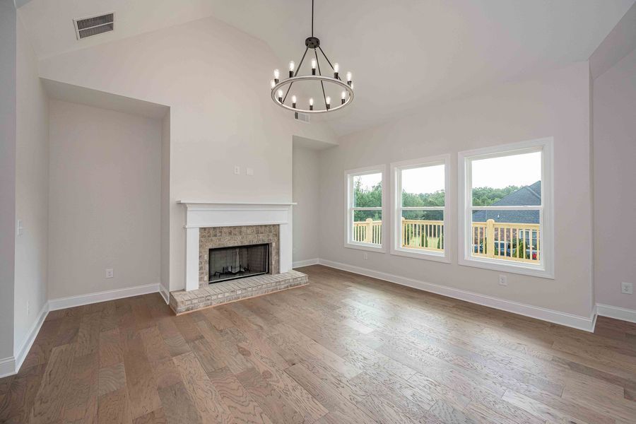 Empty living room with fireplace, wood floors, and three windows.  A chandelier hangs from vaulted ceiling.