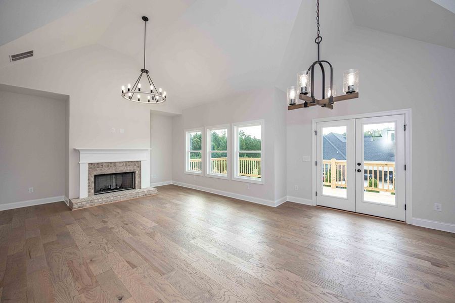 Empty living room with fireplace, windows, and French doors, hardwood floor, two chandeliers, and high vaulted ceiling.