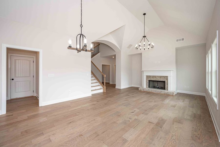 Spacious, empty living room with hardwood floors, fireplace, and chandelier. Door and stairs visible.