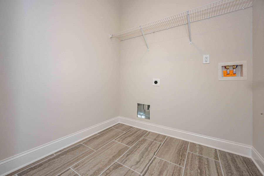 Empty laundry room with beige walls, tile floor, and wire shelf above. Dryer hookup on the right.