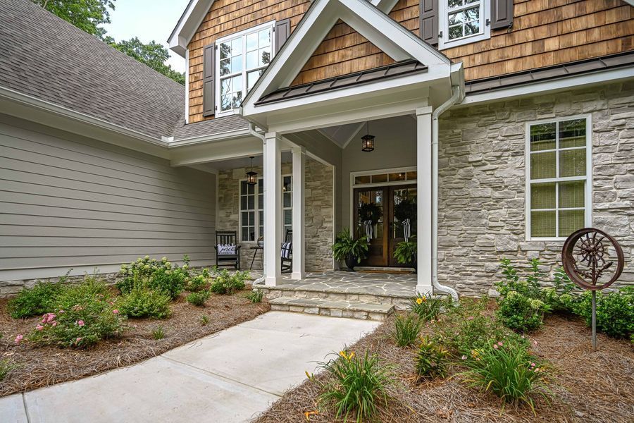 Stone-faced house with cedar shake roof, entryway with pillars, and concrete path. Landscaping with flowers.