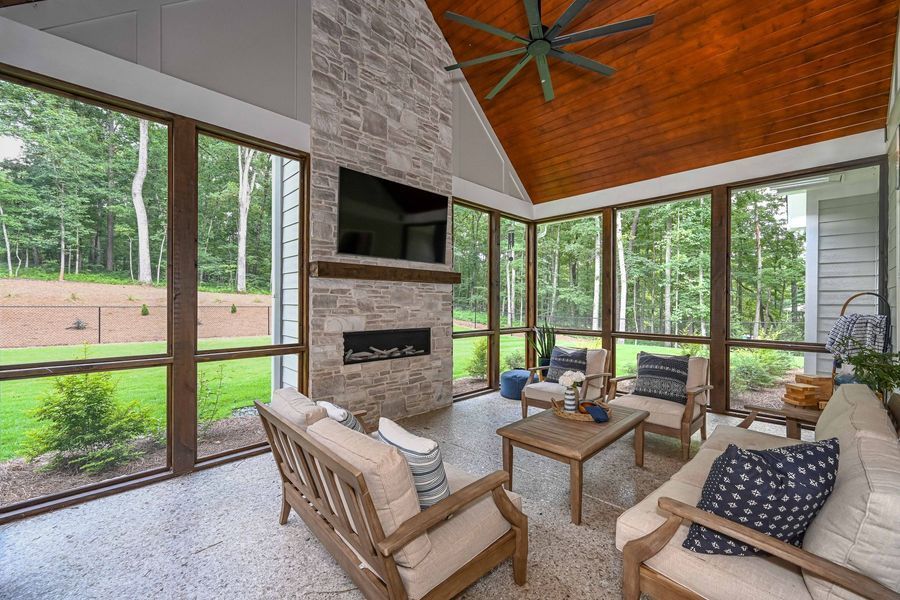 Sunroom with stone fireplace, wood ceiling, and outdoor seating overlooking a green lawn and trees.