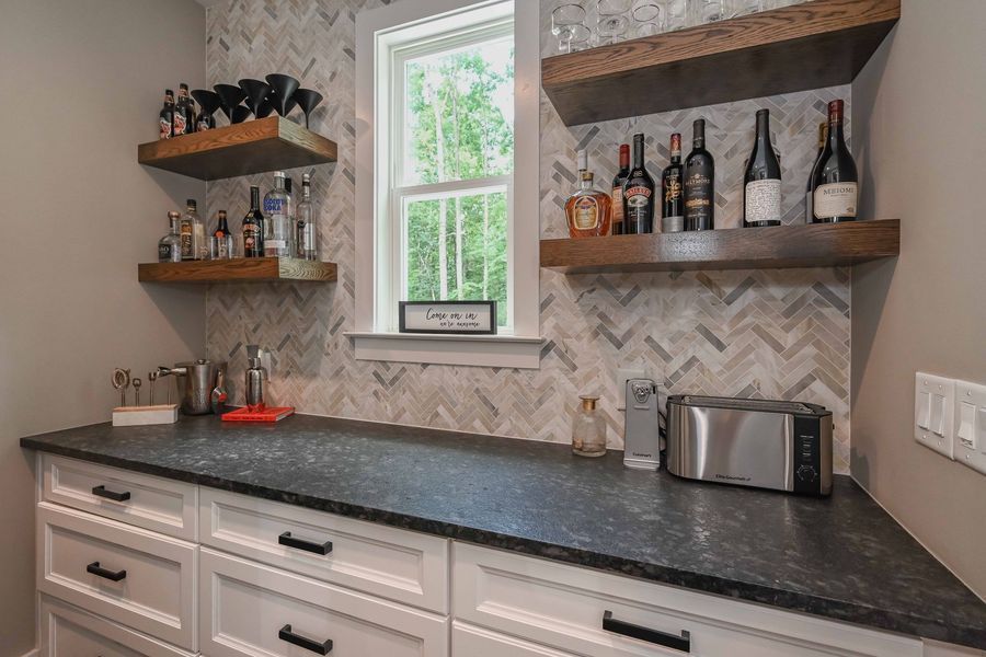 Home bar with white cabinets, black countertop, wooden shelves with liquor bottles, and a window.