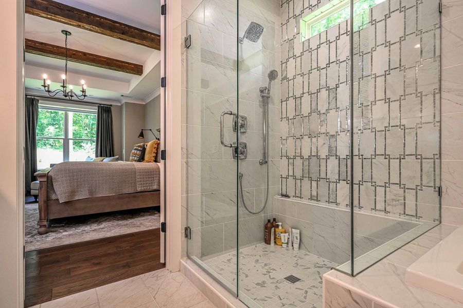 A modern bathroom with a glass shower and a peek into a bedroom, featuring marble, tile, and natural light.