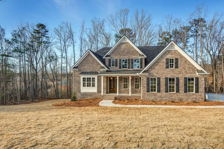 Brick house with black shutters, brown roof, on grassy lot, with trees in the background.