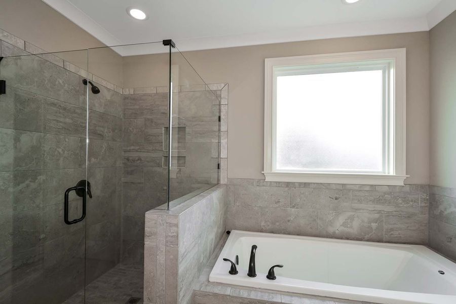 Modern bathroom with a glass shower, tub, and neutral-toned tile. White window and black fixtures.